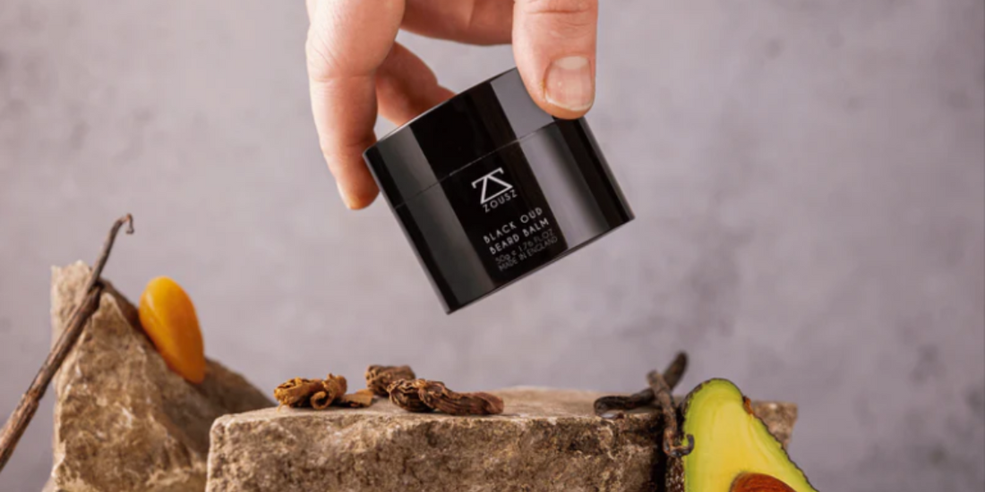 Hand holding Beard Balm above a rock and some dried fruit and an avocado.