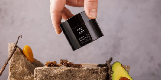 Hand holding Beard Balm above a rock and some dried fruit and an avocado.