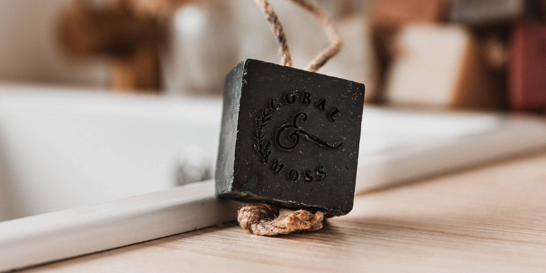 Charcoal soap on a rope sitting on edge of sink with other soaps and tap in background.