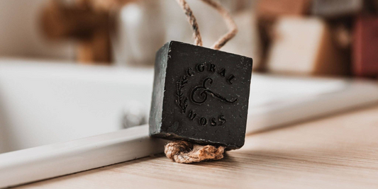 Charcoal soap on a rope sitting on edge of sink with other soaps and tap in background.