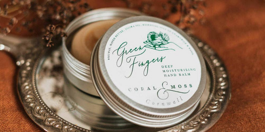 Green Finger Hand Balm open tin sitting on a hand mirror on a clothed table with herbs around it.