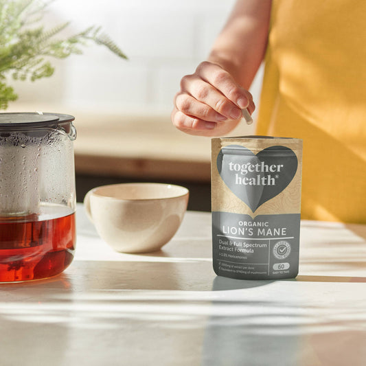 Person in the kitchen with a pot of tea and a cup. There is a plant. The hand is reaching into packet to get a tablet out.