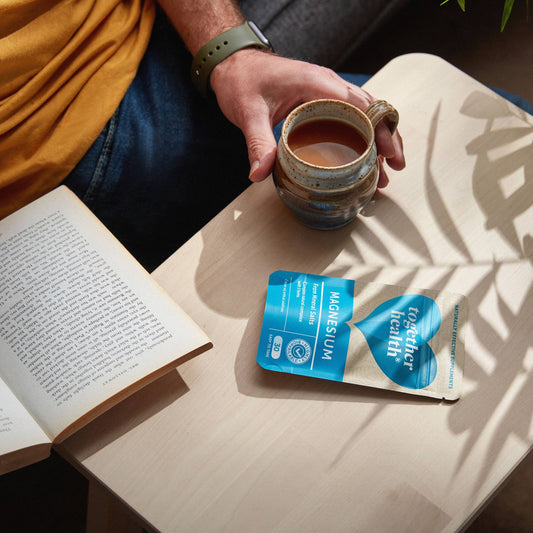 Man wearing a watch and reading a book with a coffee and a table and a packet of tablets.