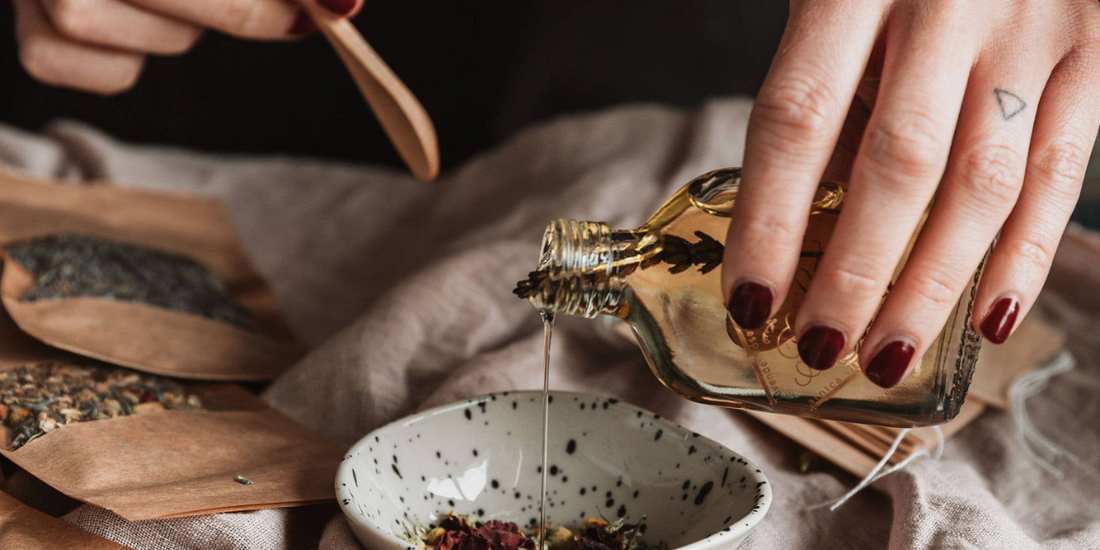 Hand pouring oil into bowl with dried flowers. Bowl is on a tablecloth with various botanicals around it.