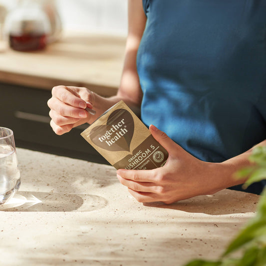 Lady holding packet of tablets in kitchen. There is also a glass of water and a plant.