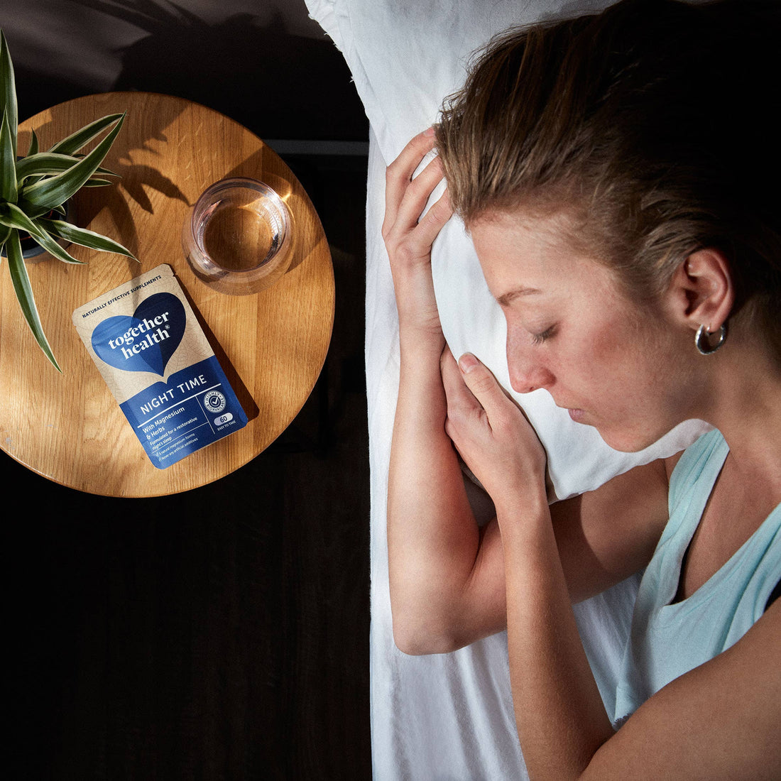 Lady sleeping on a bed with a glass of water, a plant and packet of tablets on side table next to her.
