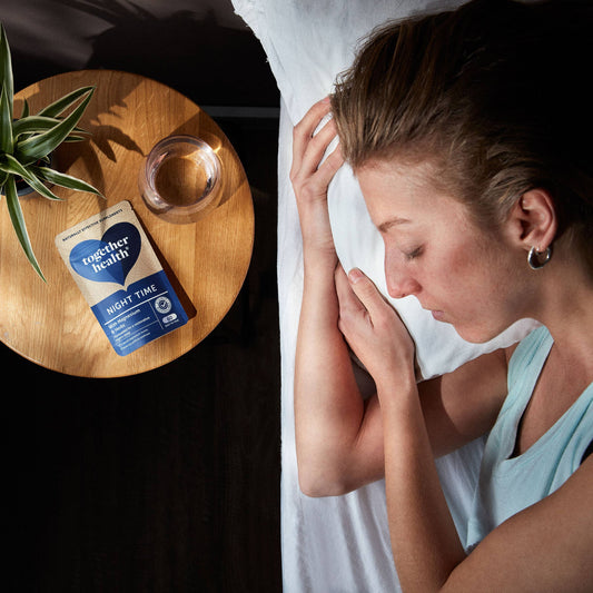 Lady sleeping on a bed with a glass of water, a plant and packet of tablets on side table next to her.