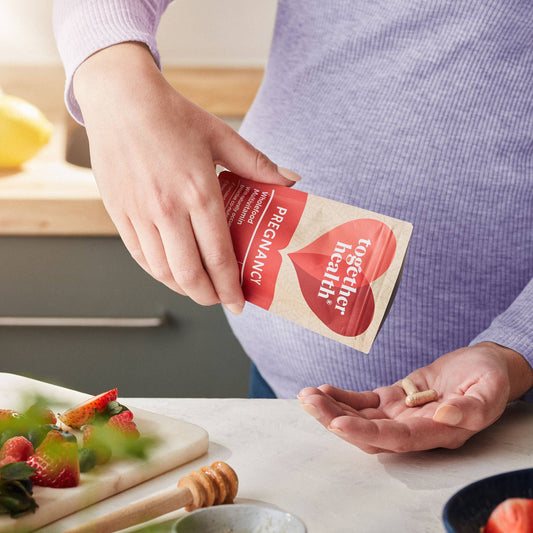 Pregnant lady shaking some tablets out to her hand in kitchen near a bench with fruit on it.