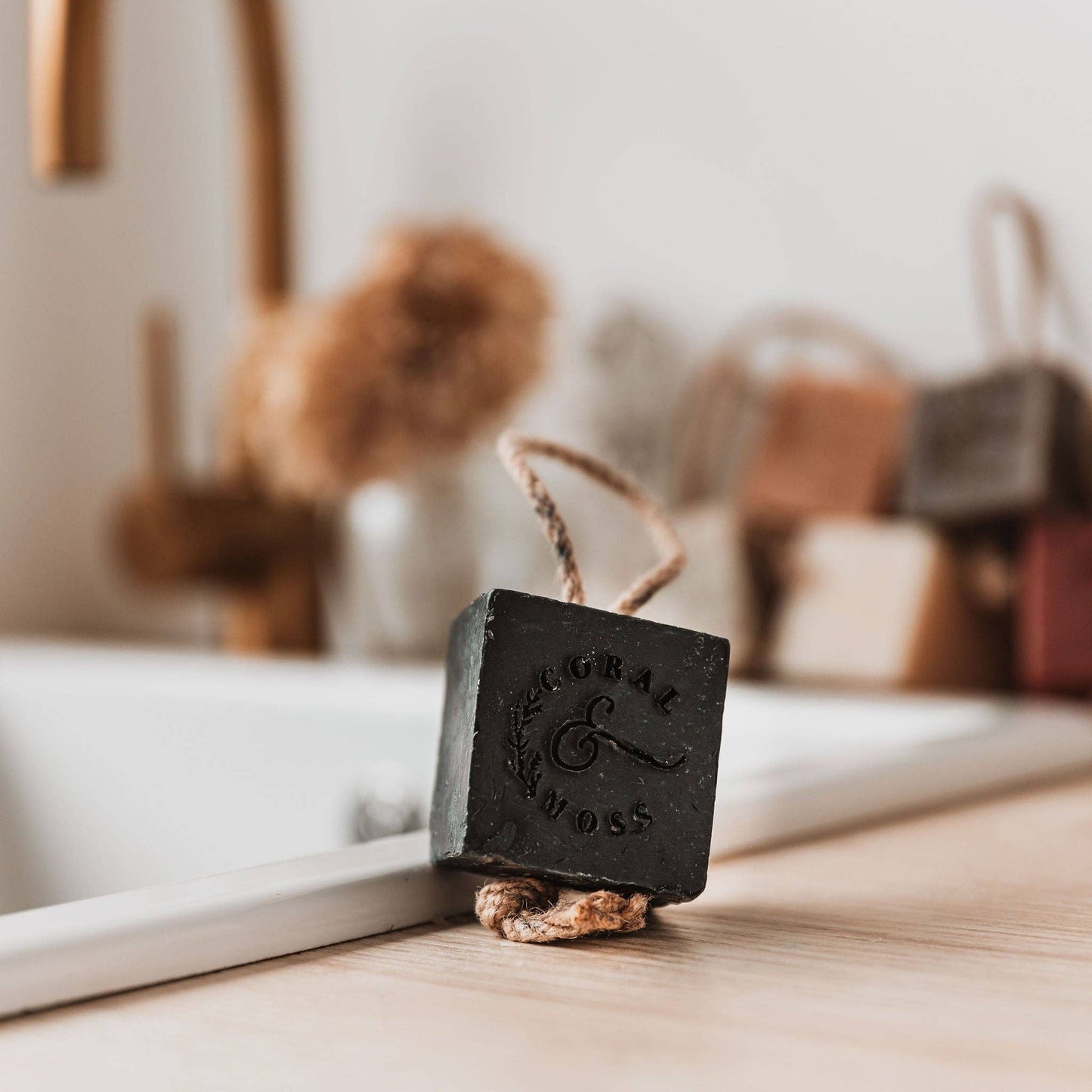 Charcoal soap on a rope sitting on edge of sink with other soaps and tap in background.
