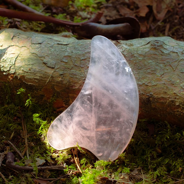 Crystal gua sha tool on a wooden log with moss and leaves in the background