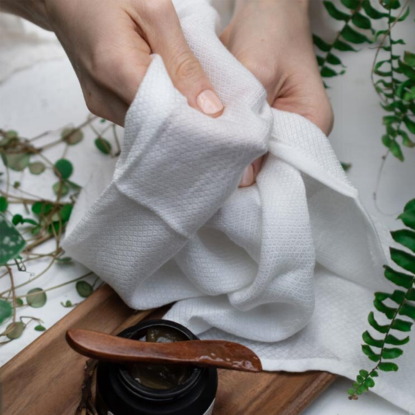 Person holding a white cloth over a wooden tray with a black container and wooden spoon, surrounded by green plants.