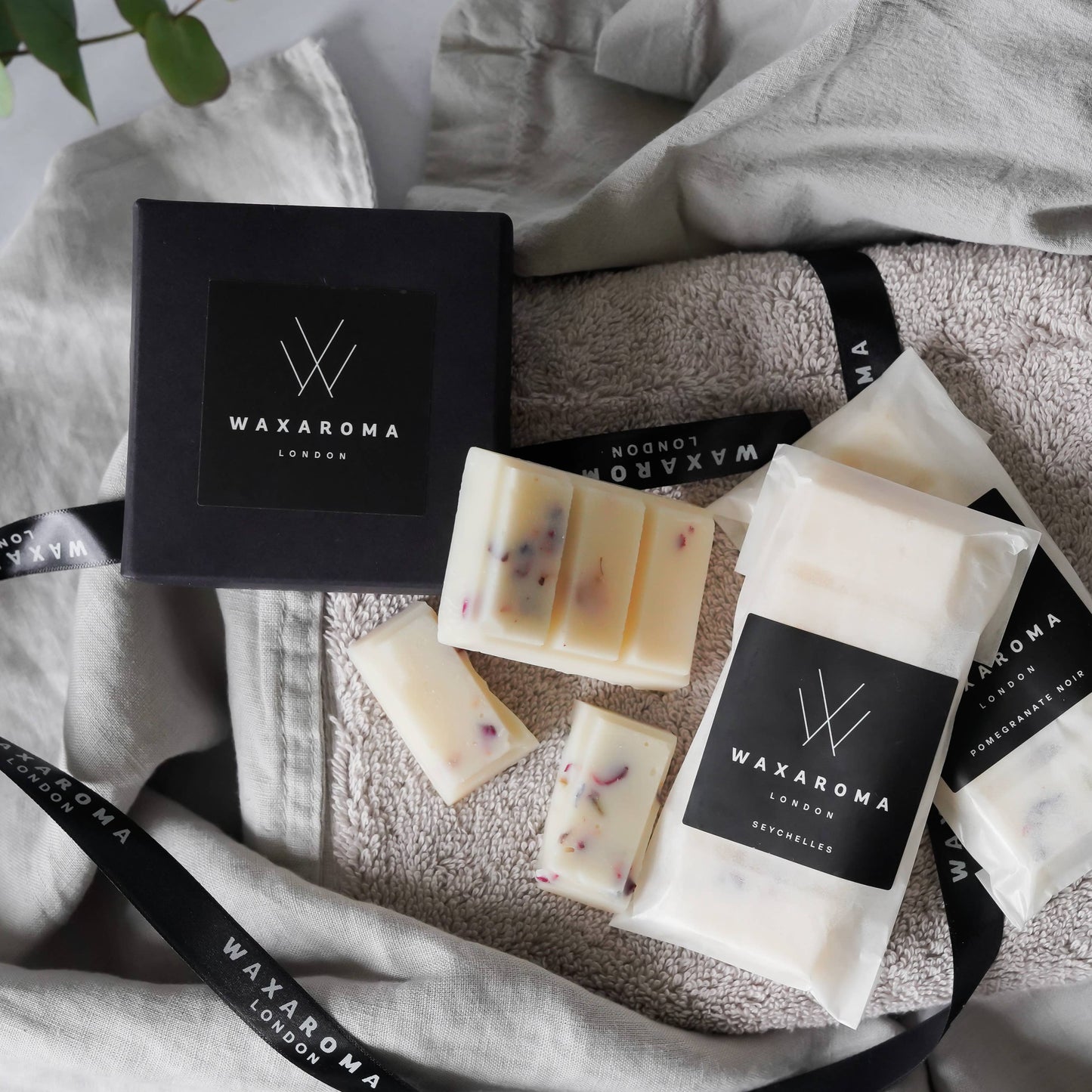 Two Waxaroma packets and one open melts bar on a towel and blanket with eucalyptus leaves and a Waxaroma branded box