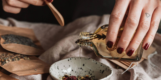 Hand pouring oil into bowl with dried flowers.  Bowl is on a tablecloth with various botanicals around it.