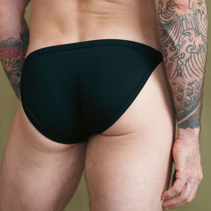 Person wearing black swim briefs with a tattooed arm on a plain background