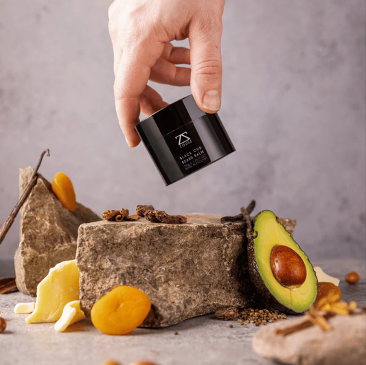 Hand holding Beard Balm above a rock and some dried fruit and an avocado.