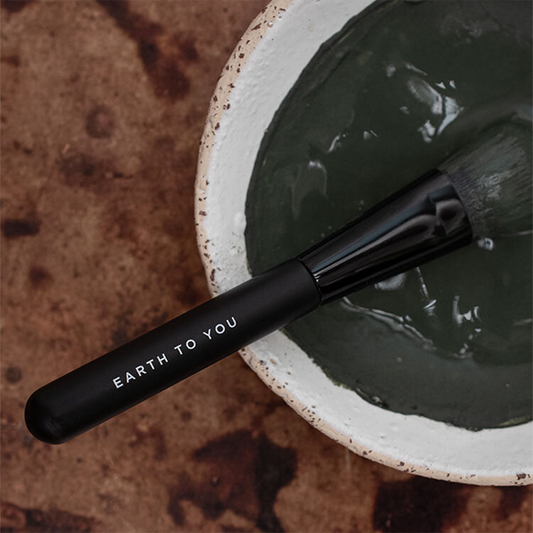 Black makeup brush with 'Earth to You' branding in a bowl of green clay on a textured surface.