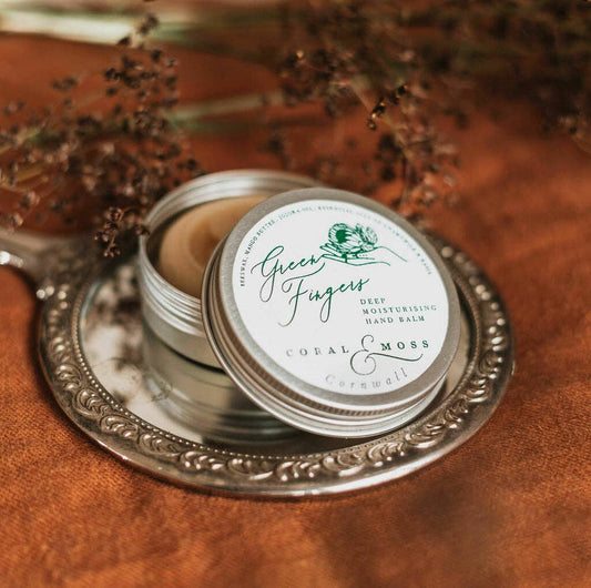 Green Finger Hand Balm open tin sitting on a hand mirror on a clothed table with herbs around it.