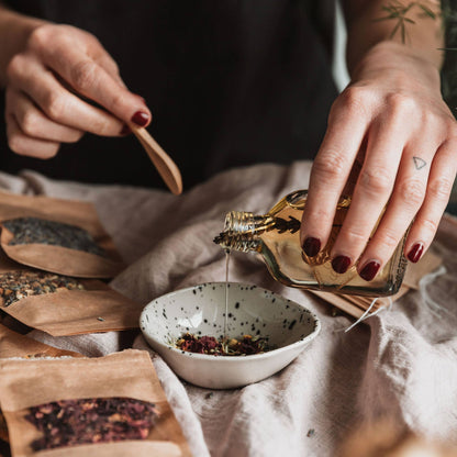 Hand pouring oil into bowl with dried flowers.  Bowl is on a tablecloth with various botanicals around it.