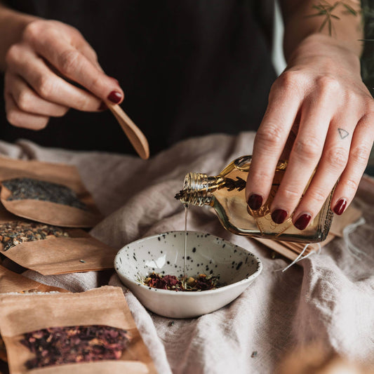Hand pouring oil into bowl with dried flowers.  Bowl is on a tablecloth with various botanicals around it.