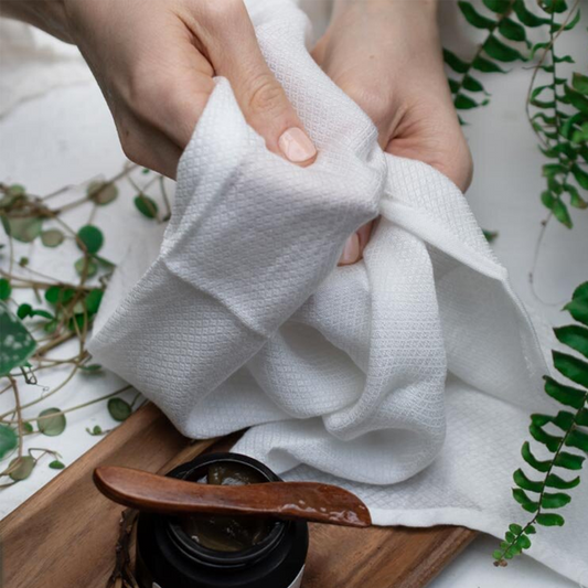 Person holding a white cloth over a wooden tray with a black container and wooden spoon, surrounded by green plants.