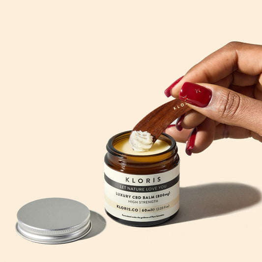 Hand using a wooden spatula to scoop out cream from a Kloris jar on a beige background